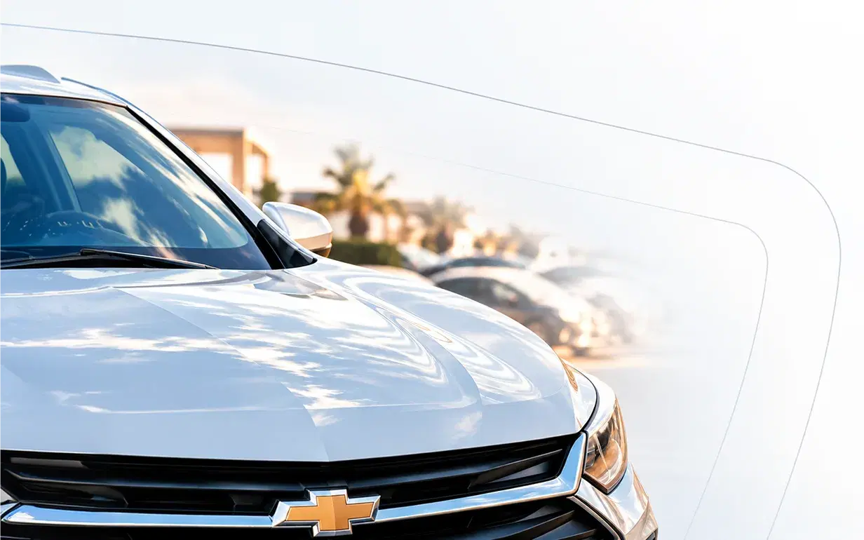 Frente de um Chevrolet prateado ao sol, com carros alinhados ao fundo